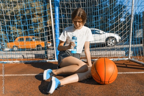 Teenager girl playing street basketball with ball on outdoor city basketball court drinking water photo