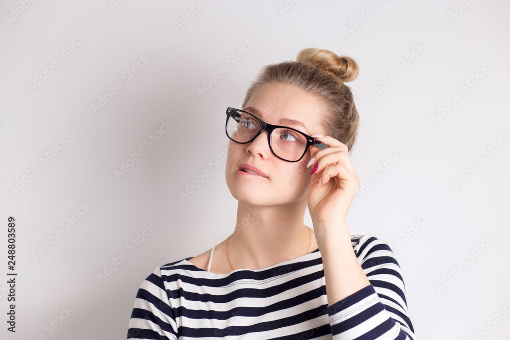 Indoor shot of cute girl looking away, having doubtful and indecisive face expression, pursuing her lips. Confused young female posing isolated at white wall