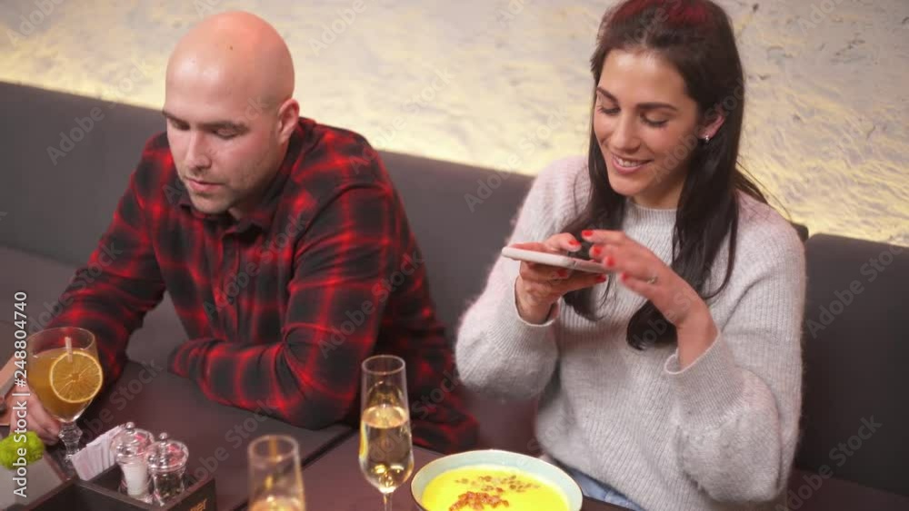 Young woman taking food pictures with smart phone in a cafe while her boyfriend is talking with a friend. Company of friends is drinking and enjoying time in a nice place. Young woman taking food pictures with smart phone in a cafe while her boyfriend is talking with a friend. Company of friends is drinking and enjoying time in a nice place.