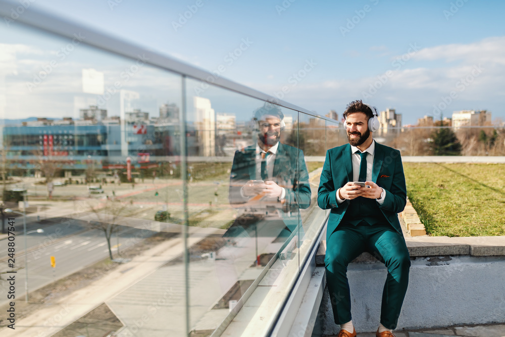 Full length of successful businessman in formal wear having headphones on ears and using smart phone while sitting on rooftop. Full length of successful businessman in formal wear having headphones on ears and using smart phone while sitting on rooftop.