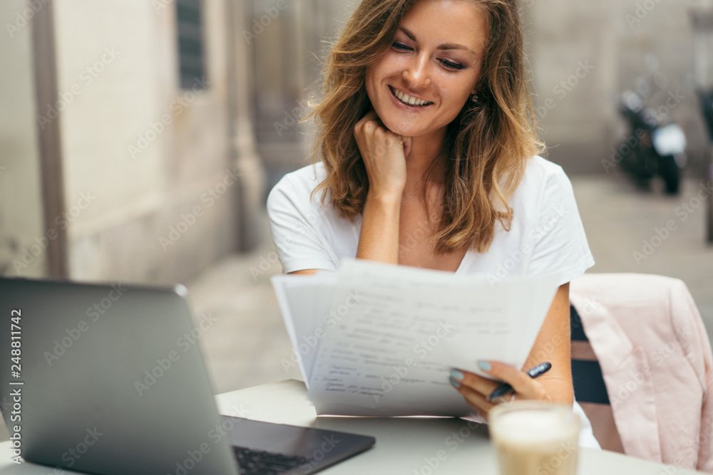 Young happy smiling girl sitting with laptop computer at desk in outdoors coffee shop and having online video lesson or doing distant job with papers, concept of freelance work or online education Young happy smiling girl sitting with laptop computer at desk in outdoors coffee shop and having online video lesson or doing distant job with papers, concept of freelance work or online education