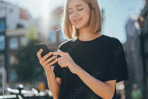 Happy smiling hipster blonde girl browsing news feed of her mobile phone while walking on the city streets and enjoying wireless connection talking with friends and using internet