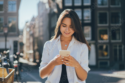 Happy smiling young girl browsing internet and typing in messenger using mobile phone while walking on the city streets and enjoying wireless connection talking with friends and using internet