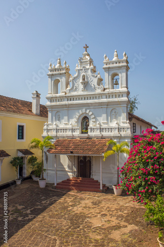 white catholic church next to the yellow house and tropical plants under the blue sky