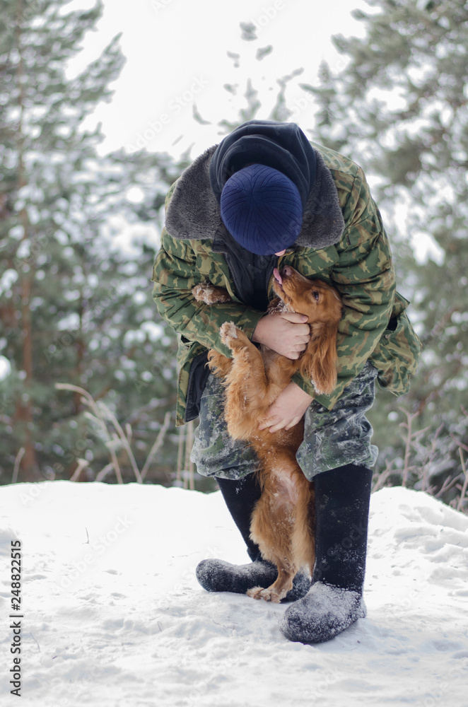 Warmly dressed man embracing loveful young russian spaniel dog in the winter forest