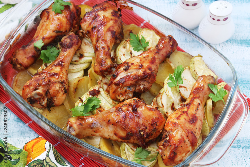 Fototapeta premium Baked chicken drumstick with potatoes and zucchini in glass form. The photo has a fork, knife and jars of salt and pepper