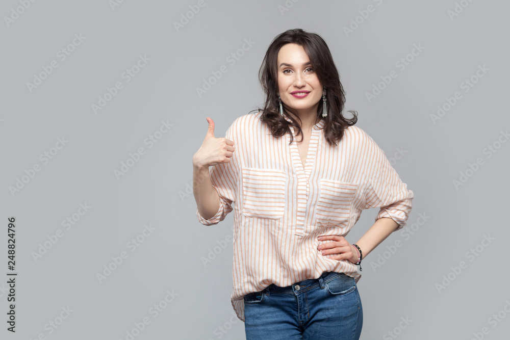 Portrait of satisfied beautiful attractive brunette woman in casual striped orange shirt with makeup and curly hairstyle standing toothy smile and thumbs up. studio shot, isolated on grey background. Portrait of satisfied beautiful attractive brunette woman in casual striped orange shirt with makeup and curly hairstyle standing toothy smile and thumbs up. studio shot, isolated on grey background.