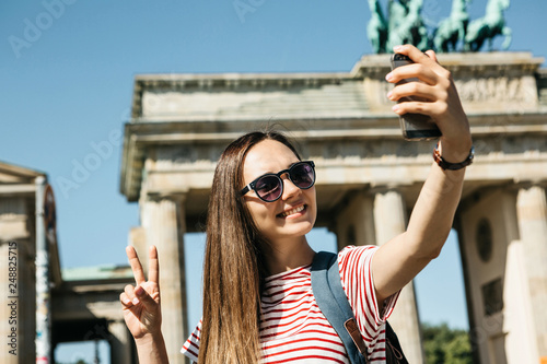 Young beautiful positive girl makes selfie against the background of the Brandenburg Gate in Berlin in Germany or takes pictures of sights.
