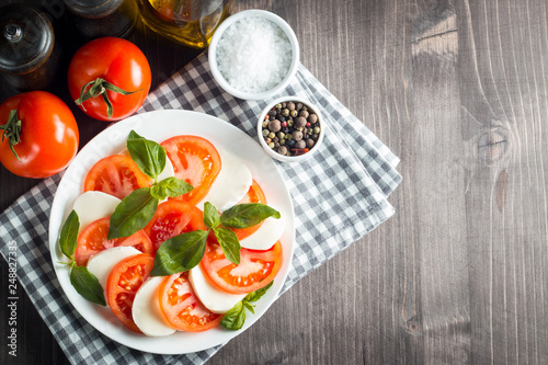 Photo of Caprese Salad with tomatoes, basil, mozzarella, olives and olive oil on wooden background. Italian traditional caprese salad ingredients. Mediterranean, organic and natural food concept.