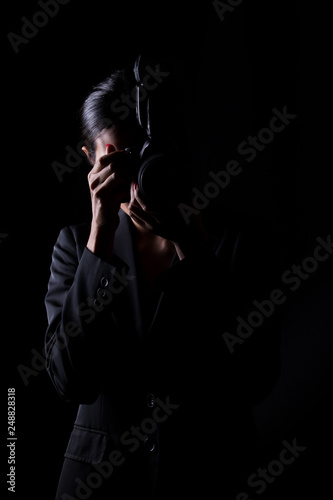 Asian Woman Photographer hold camera with external flash point to shoot subject, wear gray suit. studio lighting black background isolated low key exposure, reporter journalist take photo celebrity