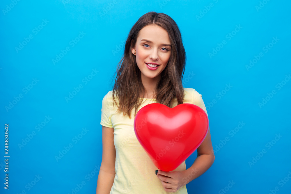 valentine's day and people concept - smiling young woman or teenage girl in yellow t-shirt with red heart-shaped balloon over bright blue background