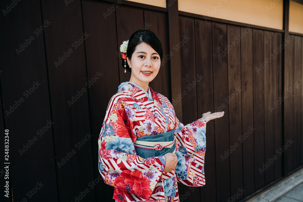 japanese local girl in floral kimono smiling face camera with welcome hand gesture standing on wooden wall in background. lady in traditional dress host telling people to come to visit kyoto japan.