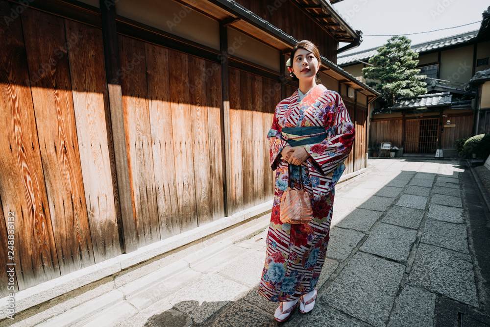 full length of young japanese woman in traditional cloth going to visit family in old town in new year walking along the wooden wall on stone road in ishibe alley. local girl in kimono enjoy sunlight