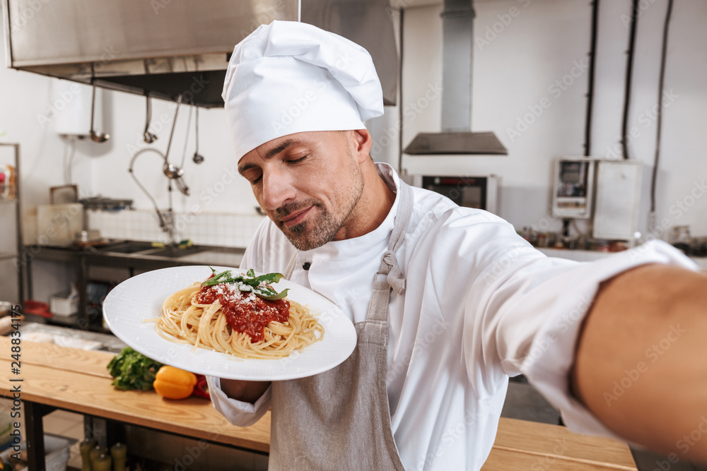 Image of professional man chief in apron taking selfie, while standing at kitchen in restaurant Image of professional man chief in apron taking selfie, while standing at kitchen in restaurant