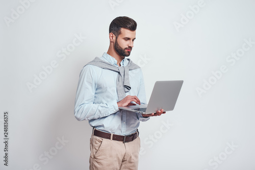 Modern technologies. Successful young man with a beard using his laptop while standing against grey background.