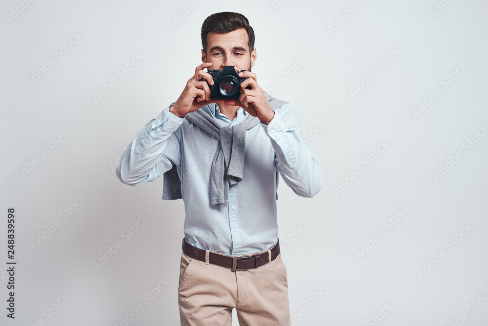 Smile! Close up photo of handsome male photographer in casual clothes who is taking photos with camera on the grey background Smile! Close up photo of handsome male photographer in casual clothes who is taking photos with camera on the grey background