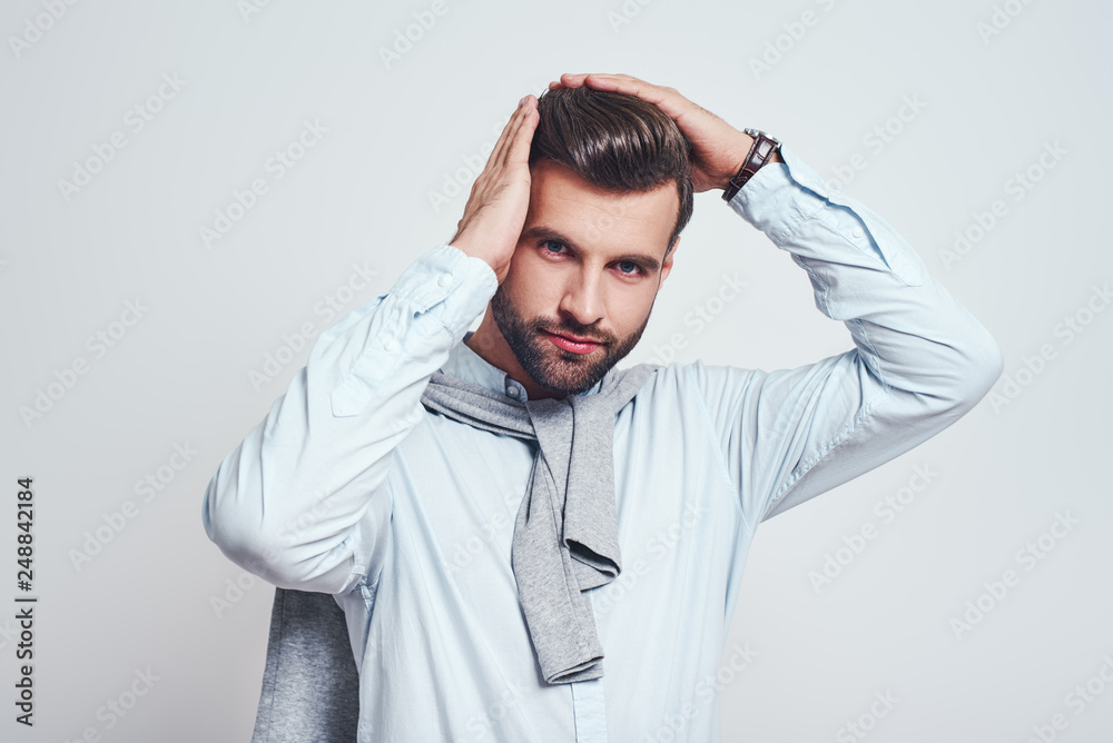 Everything must be perfect. Close up portrait of handsome young man keeping hands on hair and looking ar camera while standing against grey background Everything must be perfect. Close up portrait of handsome young man keeping hands on hair and looking ar camera while standing against grey background