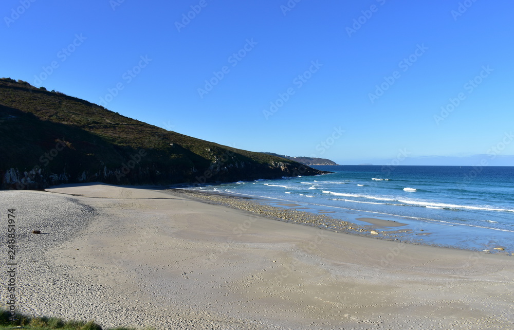 Beach with morning light. Golden sand, rocks and blue sea with waves and foam. Clear sky, sunny day. Galicia, Spain.