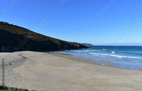 Beach with morning light. Golden sand, rocks and blue sea with waves and foam. Clear sky, sunny day. Galicia, Spain.