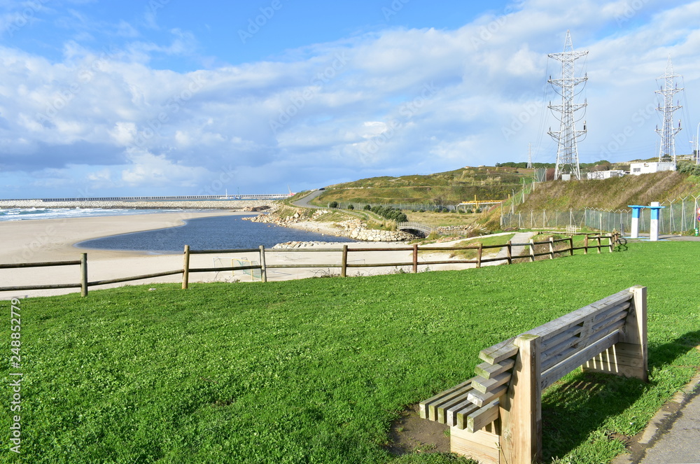 Beach with grass, wooden bench and fence. Industrial port and blue river, cloudy sky. Sabon, Galicia, Spain.
