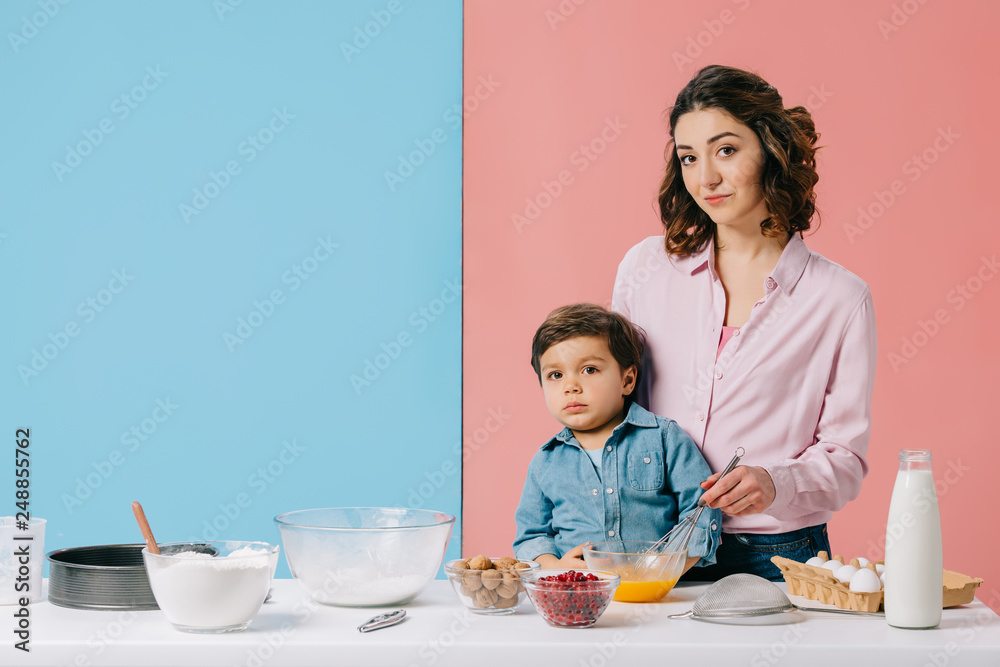 mother with adorable son whipping eggs at white kitchen table with baking ingredients on bicolor background mother with adorable son whipping eggs at white kitchen table with baking ingredients on bicolor background