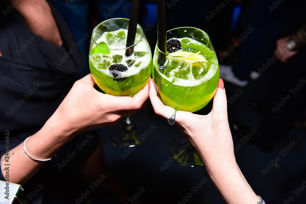 Cheers (cin cin) with two green cocktails with a black raspberry. Women hands