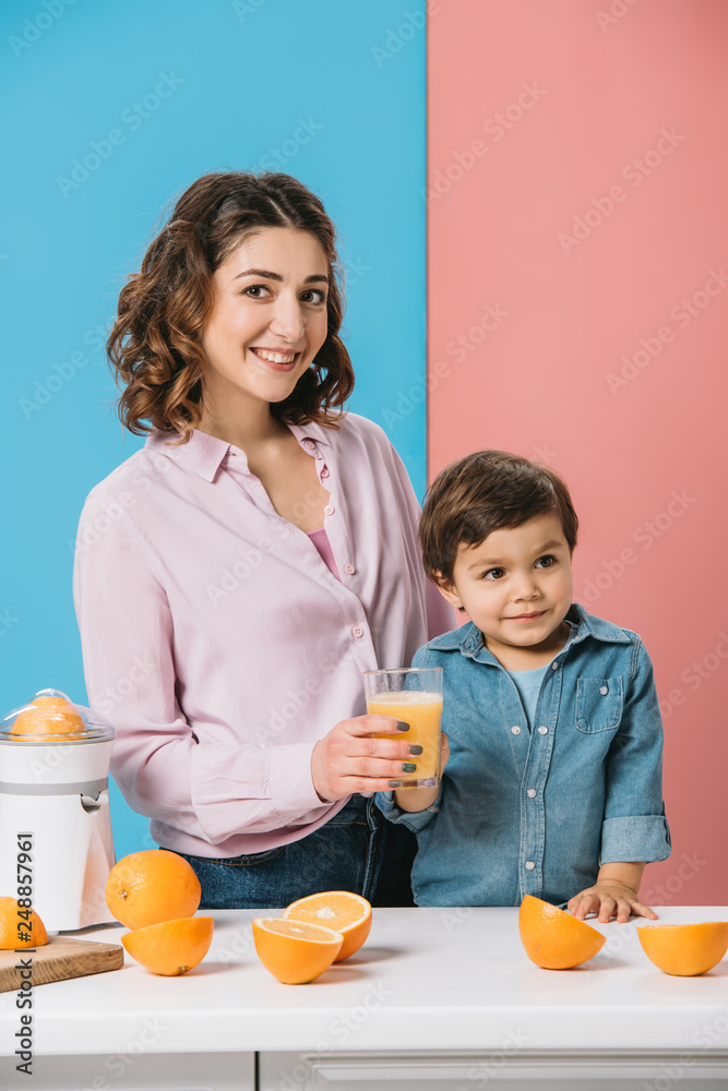 happy mother giving full glass of fresh orange juice to adorable smiling little son on bicolor background