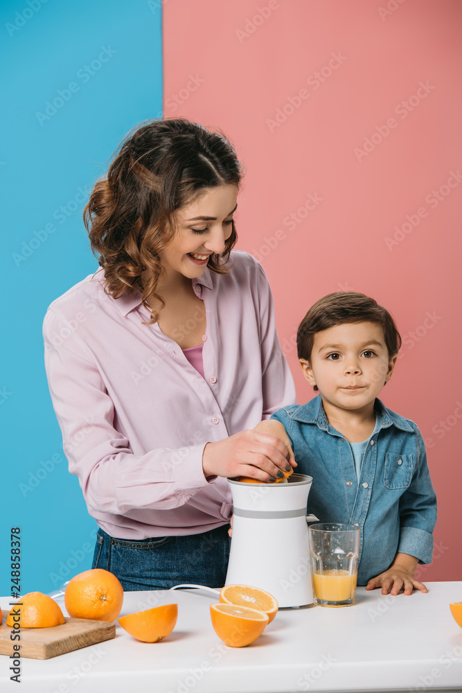 smiling mother and adorable little son squeezing fresh orange juice together on bicolor background smiling mother and adorable little son squeezing fresh orange juice together on bicolor background