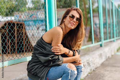 Fashion portrait of trendy young woman wearing sunglasses, jeans with halls and bomber jacket in the city