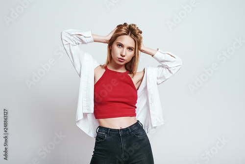 Feeling beautiful. Pretty blonde woman in red tank top keeping hands in hair and looking at camera while standing against grey background