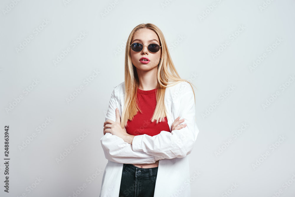 Beauty in everything. Young and stylish blonde woman in sunglasses with crossed arms standing against grey background. Fashion look Beauty in everything. Young and stylish blonde woman in sunglasses with crossed arms standing against grey background. Fashion look