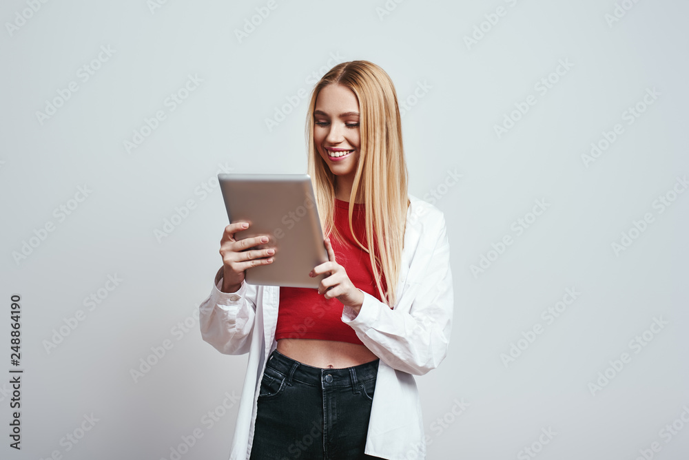 Chatting with friends. Cute blonde woman in a casual wear is using her digital tablet and smiling while standing on a grey background. Chatting with friends. Cute blonde woman in a casual wear is using her digital tablet and smiling while standing on a grey background.