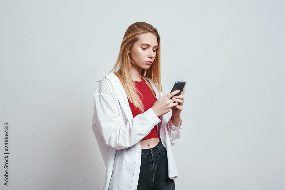 Always available. Young blonde woman in a casual wear using her smart phone while standing in studio on a grey background. Digital concept Always available. Young blonde woman in a casual wear using her smart phone while standing in studio on a grey background. Digital concept