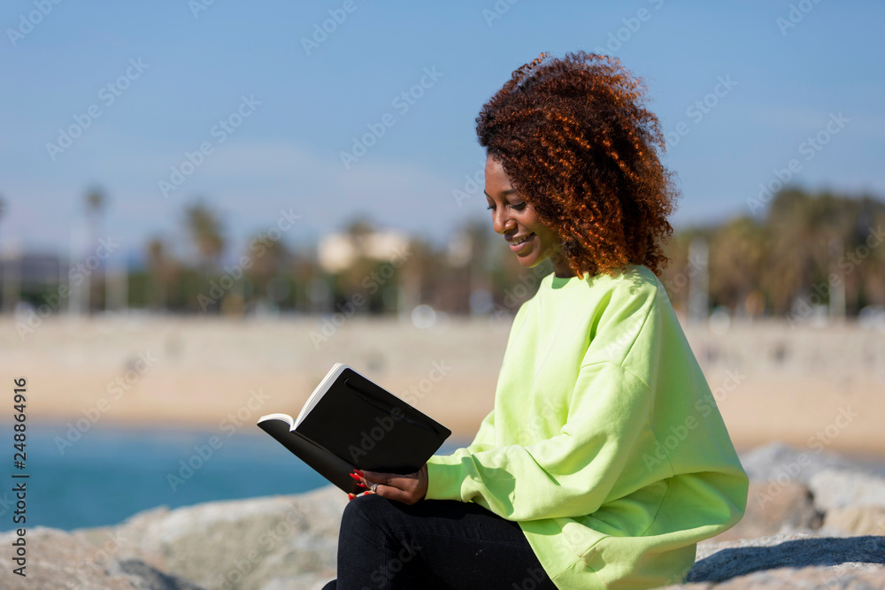 Side view of young curly afro woman sitting on a breakwater holding a book while smiling and reading outdoors