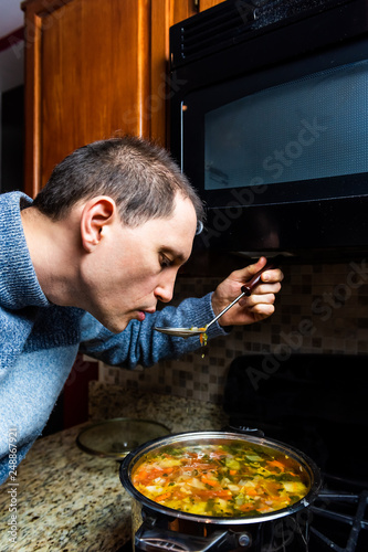 Closeup vertical portrait of young man cooking tasting homemade hot winter vegetable soup with ladle in cold sweater