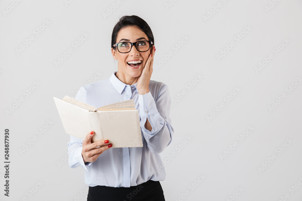 Photo of satisfied female worker wearing eyeglasses reading book in the office, isolated over white background