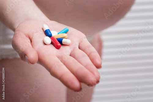 Pregnant woman in lingerie holding multicolored pills on palm on light background. Close-up
