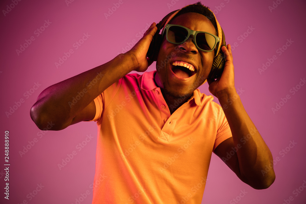 Fototapeta premium The young handsome happy hipster man listening music with headphones at black studio with neon lights. Disco, night club, hip hop style, positive emotions, face expression, dancing concept