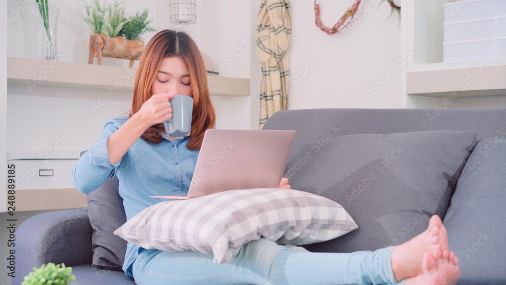 Portrait of beautiful attractive Asian woman using computer or laptop holding a warm cup of coffee or tea while lying on the sofa when relax in living room at home. Lifestyle women at home concept.