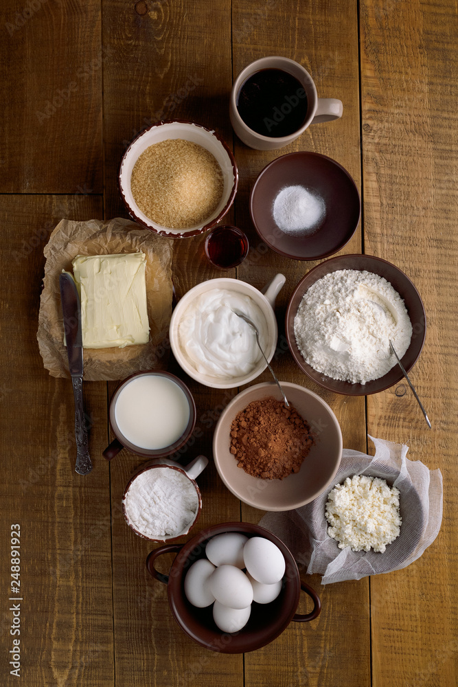 Different ingredients for baking - milk, butter, eggs and flour, cottage cheese, sour cream, brown sugar, cocoa, butter on rustic background, top view