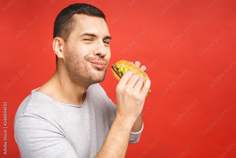 Young man holding a piece of hamburger. Student eats fast food. Burger is not helpful food. Very hungry guy. Diet concept. Isolated over red background. Young man holding a piece of hamburger. Student eats fast food. Burger is not helpful food. Very hungry guy. Diet concept. Isolated over red background.