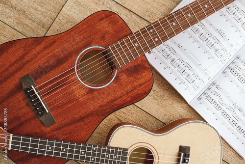 Music education. Brown acoustic guitar and ukulele lying on the wooden floor with music notes. Top view