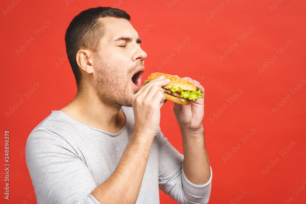 Young man holding a piece of hamburger. Student eats fast food. Burger is not helpful food. Very hungry guy. Diet concept. Isolated over red background. Young man holding a piece of hamburger. Student eats fast food. Burger is not helpful food. Very hungry guy. Diet concept. Isolated over red background.