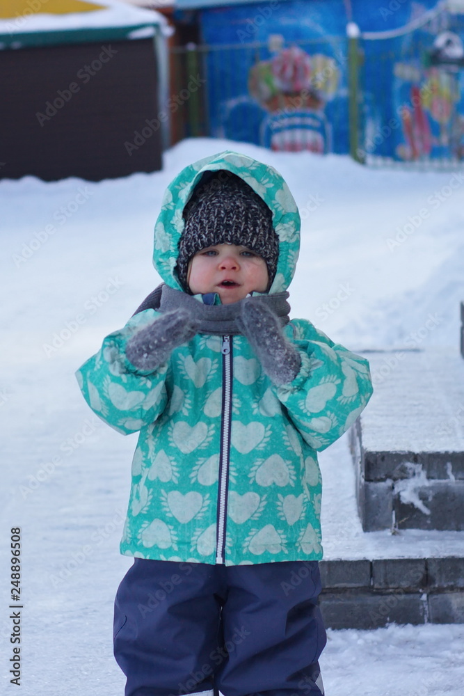 A blue-eyed little girl in a knitted hat, scarf, mittens and a green winter jacket claps her hands. Snow background. Close up.