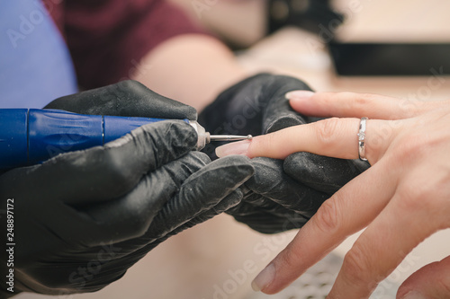 A manicurist in black latex gloves makes a hardware manicure to a client using a manual electric milling machine in a beauty salon, close up.