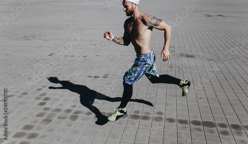 Athletic man with a naked torso with tattoos and headband on his head dressed in the black leggings and blue shorts runs on paving slabs on a warm sunny day