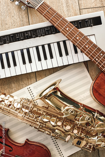 Vertical top view of different musical instruments: synthesizer, guitar, saxophone and violin lying on the sheets for music notes over wooden floor