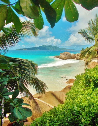 View of turquoise ocean with tropical beach, green vegetation and rocks near the shore. Through green leaves or foliage is an unsharp island visible in the distance. Summer holidays on the Seychelles