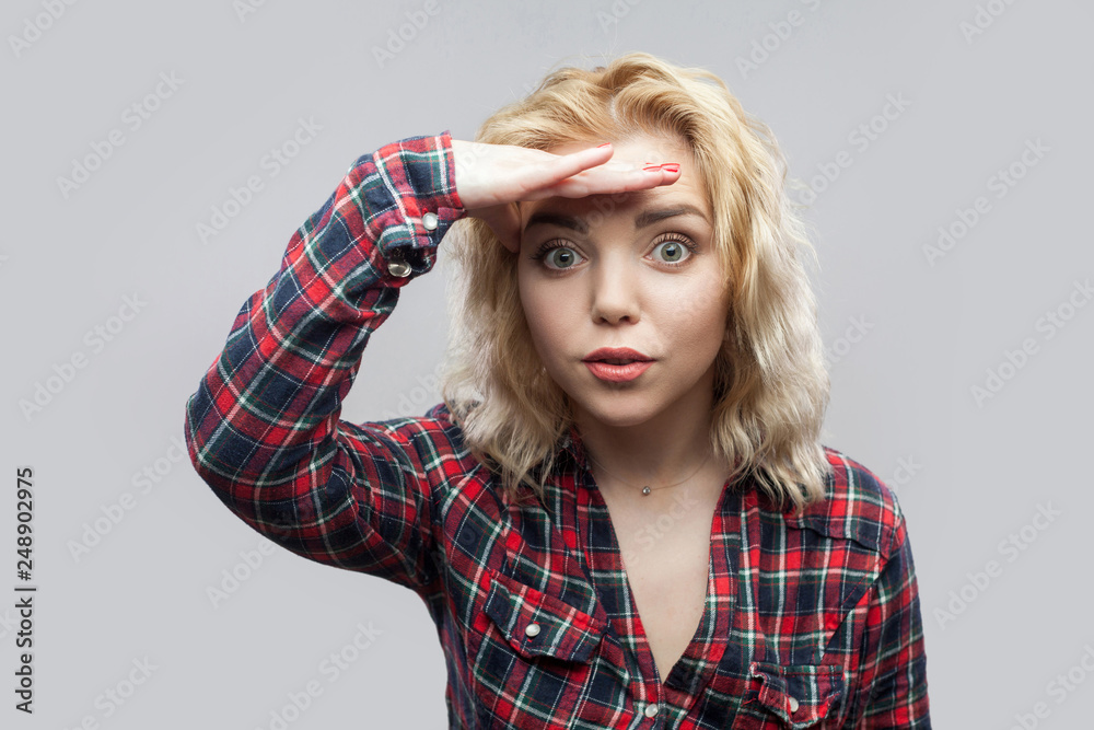 Portrait of surprised beautiful blonde young woman in casual red checkered shirt standing and looking at camera with hand on forhead. indoor studio shot, isolated on grey background.