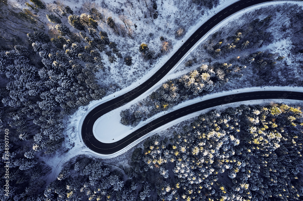 Aerial drone view of a curved winding road through the forest high up in the mountains in the winter with snow covered trees and curved streets in winter